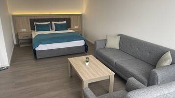 A modern hotel room with a bed, a grey sofa, a wooden coffee table, and a nightstand.