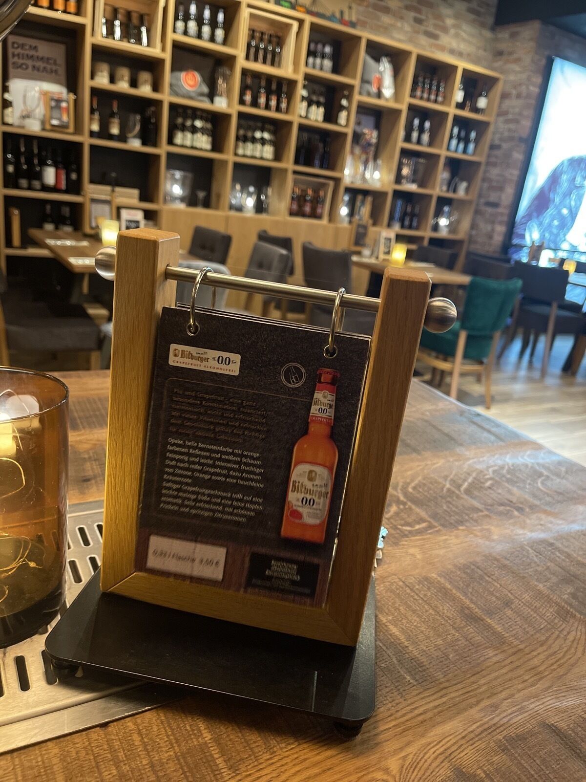 Un présentoir en bois sur lequel est posée une bouteille de bière Bitburger 0.0, entouré d’étagères remplies de diverses bouteilles et de verres, dans un décor de bar.