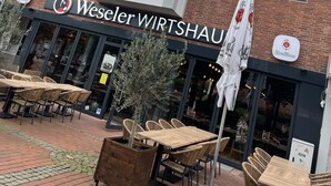 Un edificio in mattoni di due piani con un ristorante chiamato "Weseler WIRTSHAU" al piano terra. Sono presenti tavoli e sedie all'aperto, e un ombrellone grande.