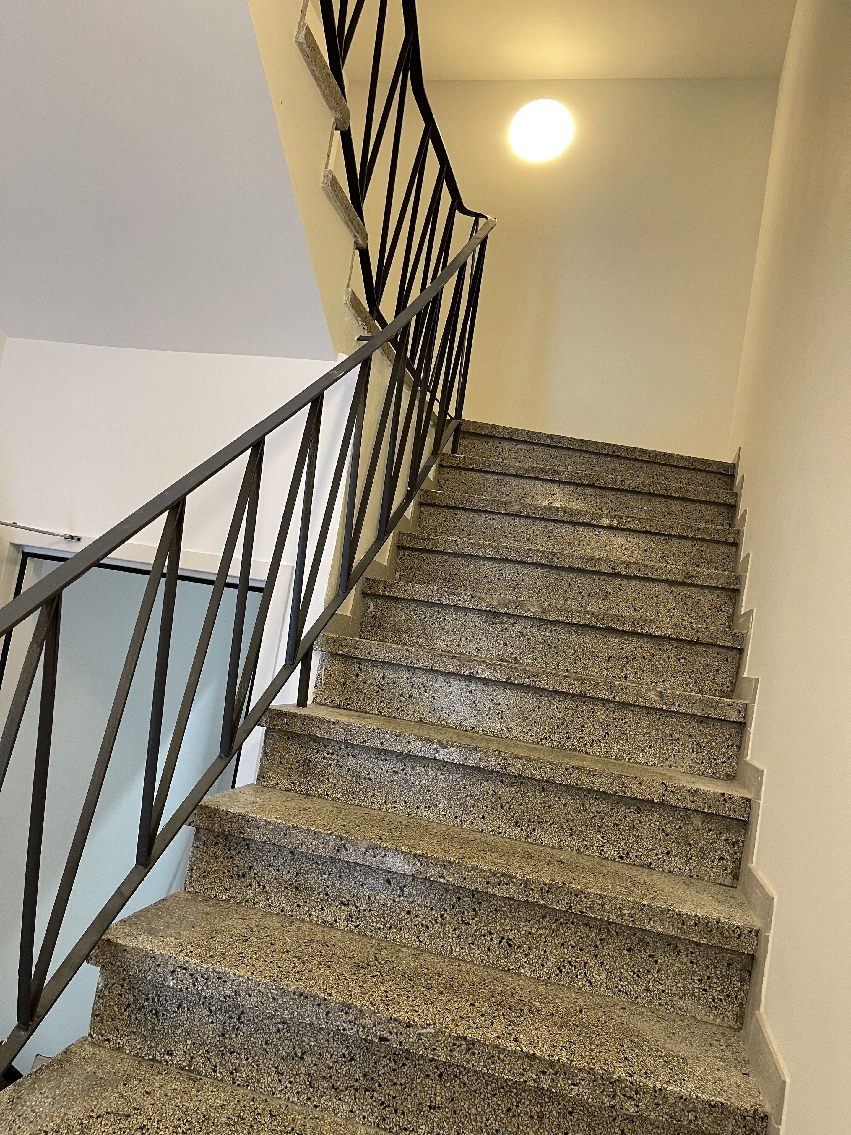 Una scala con gradini in granito e ringhiera in metallo nero, che conduce a un pianerottolo con un lampadario.