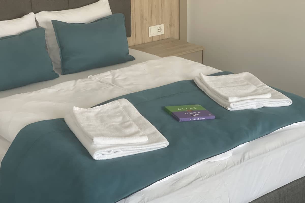 A neatly made bed with white linens, teal pillows, and a teal bedspread. A folded white towel and a book titled 'ALLES GUTE' are placed on the bed.