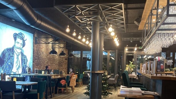 Interno di un ristorante con pavimento in legno, lampadine pendenti, un'area bar e un grande murale sulla parete.