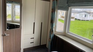 A bedroom with a bed, a wardrobe, a window with curtains, and a view of a grassy area with mobile homes. - Premium 2 bed Caravan Sleeps 6 With Huge Decking (Clacton-on-Sea)