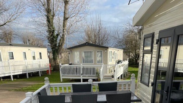 A covered outdoor deck with a table and chairs, a white railing, and a view of a house in the background. - Premium 2 bed Caravan Sleeps 6 With Huge Decking (Clacton-on-Sea)