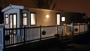 A single-story mobile home with a fenced deck, a small porch, and a visible door. - Premium 2 bed Caravan Sleeps 6 With Huge Decking (Clacton-on-Sea)