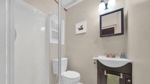 Un baño con inodoro blanco, cabina de ducha de vidrio, tocador oscuro con una toalla colgada y un cuadro en la pared.