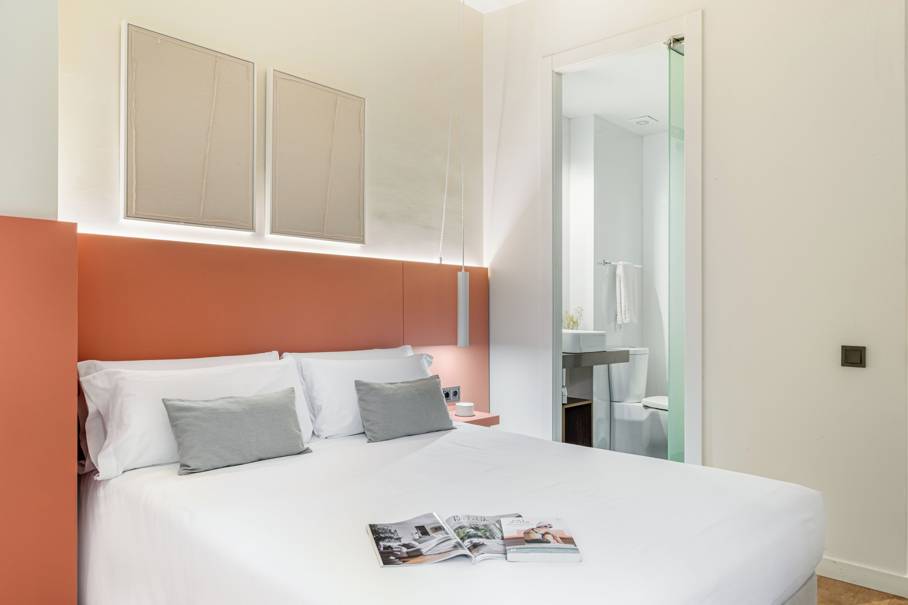 Een moderne slaapkamer met een groot bed, een nachtkastje, een tijdschrift, en een zichtbare badkamer door een open deur.