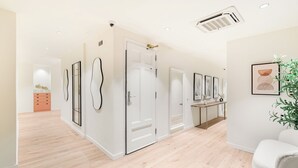 A hallway with wooden flooring, white walls, and a white door. There are framed artworks on the walls, a small plant, and a white chair. - LeMar by Aspasios (Barcelona)