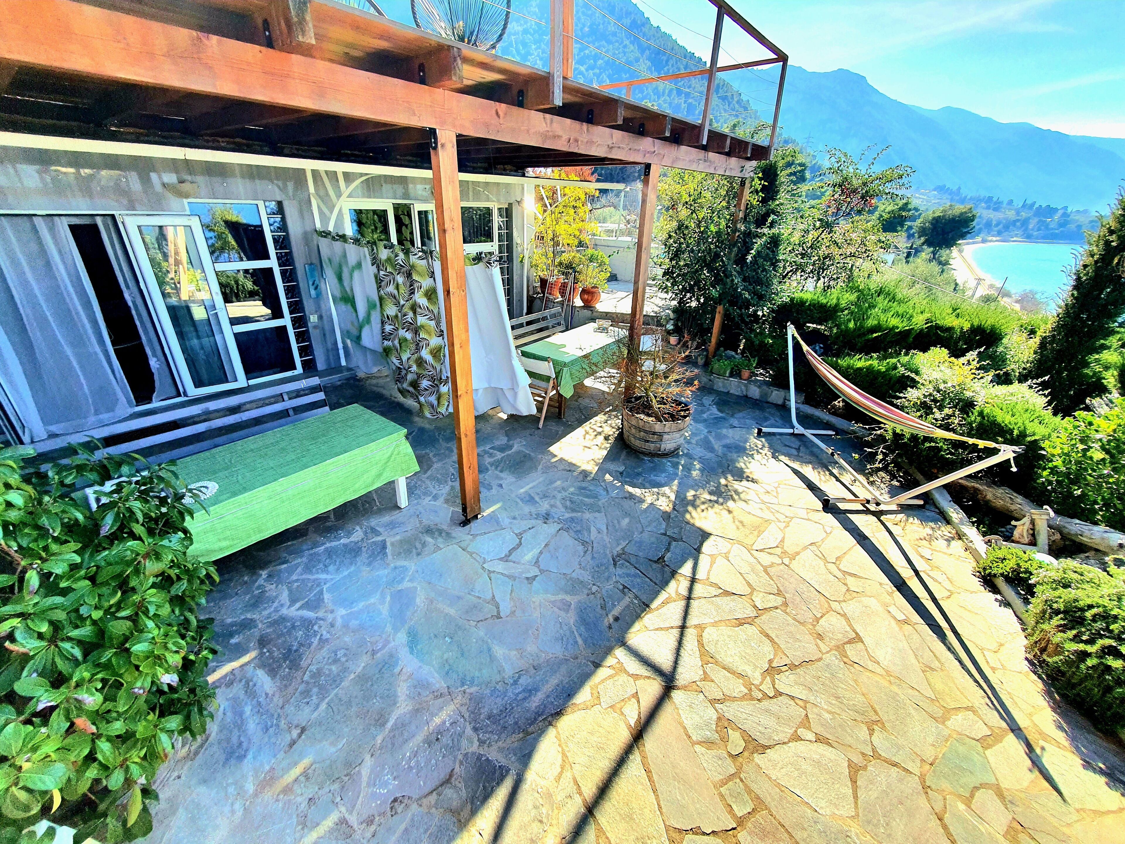 A stone patio with a hammock, a wooden pergola, and a view of a beach and mountains.