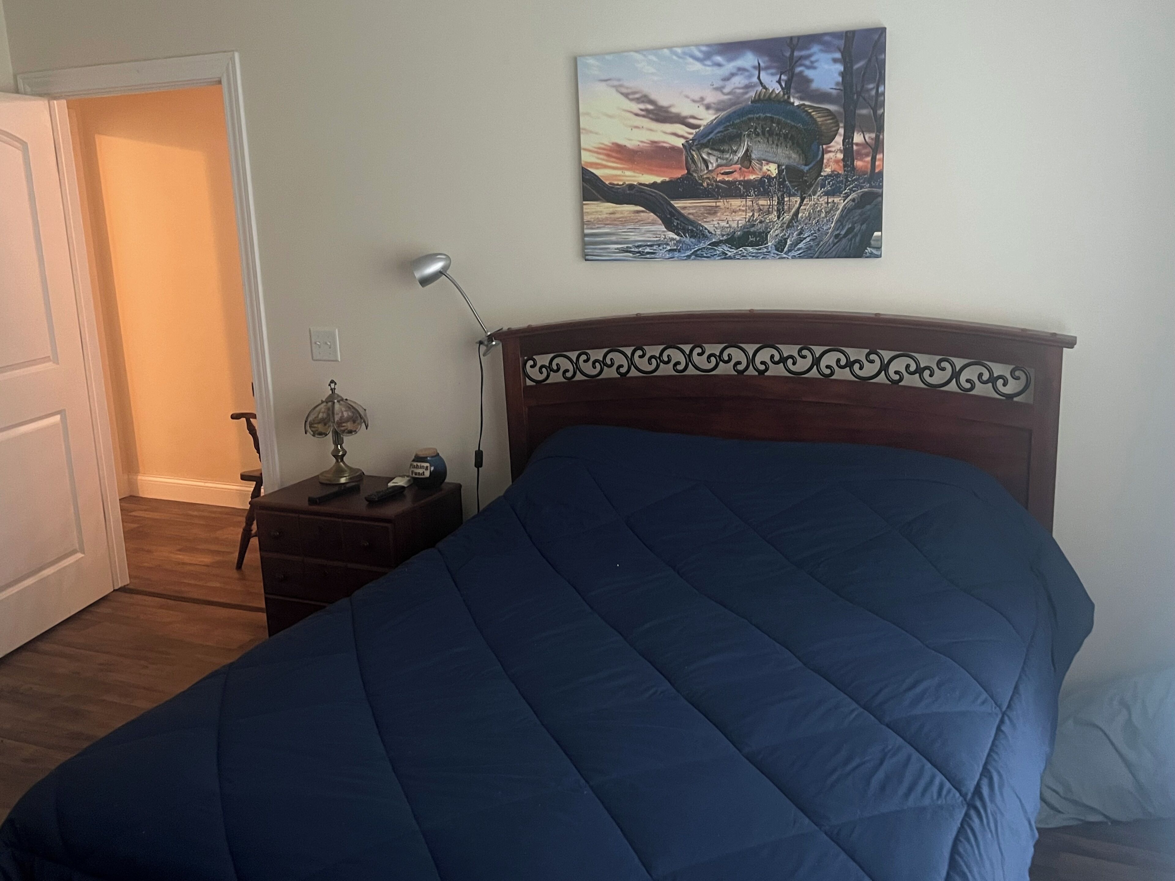 A bedroom with a large bed, a nightstand with a lamp, a painting on the wall, and a door leading to another room.