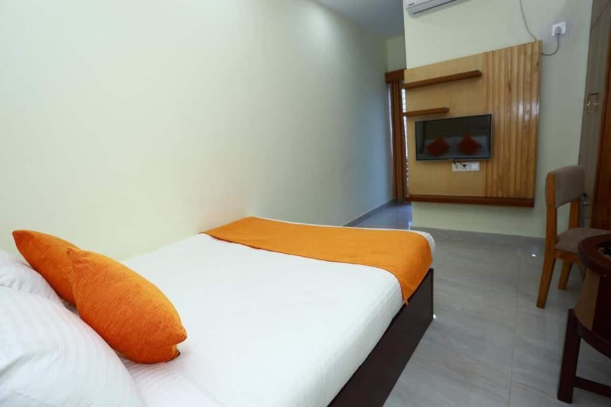 Una cama individual con un almohadón naranja y una colcha blanca, una silla de madera y un televisor fijado a un panel de pared de madera.