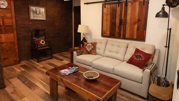 Un salón acogedor con una mesa de centro de madera, un sofá color crema con cojines estampados, una cesta de mimbre y un revestimiento de madera rústica.