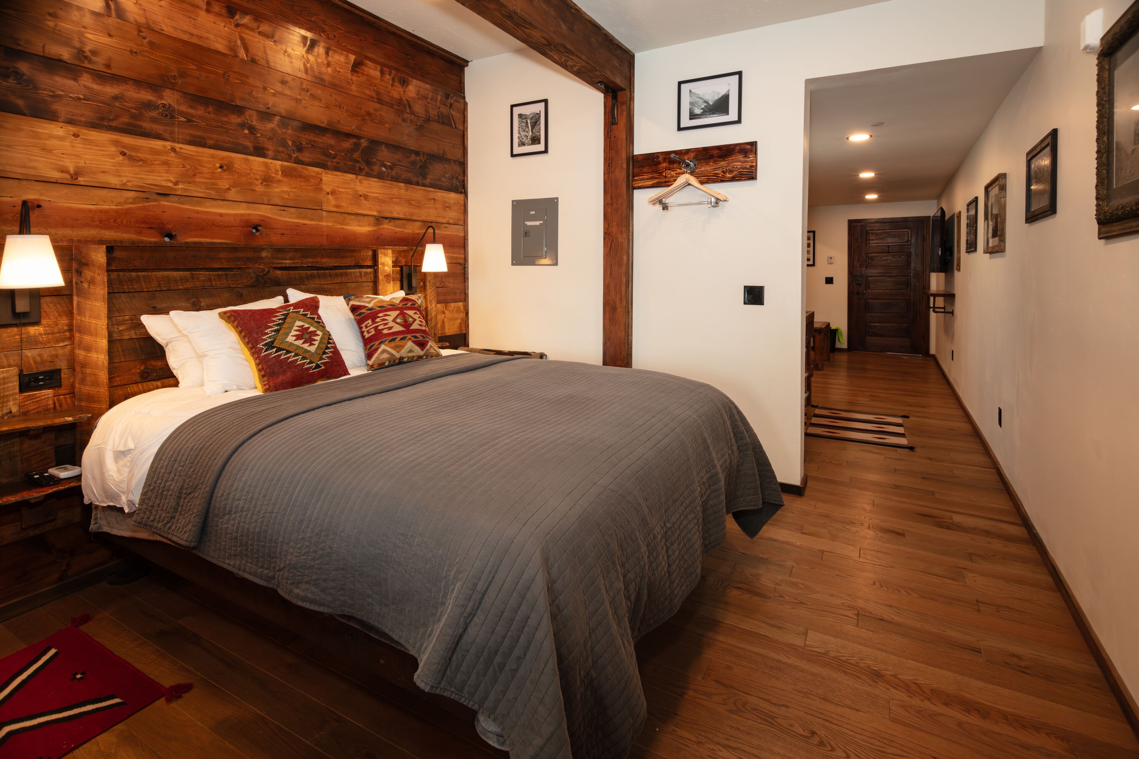 Une chambre à coucher avec une tête de lit en bois, un grand lit recouvert d’une couverture grise, et un couloir menant à une autre pièce.