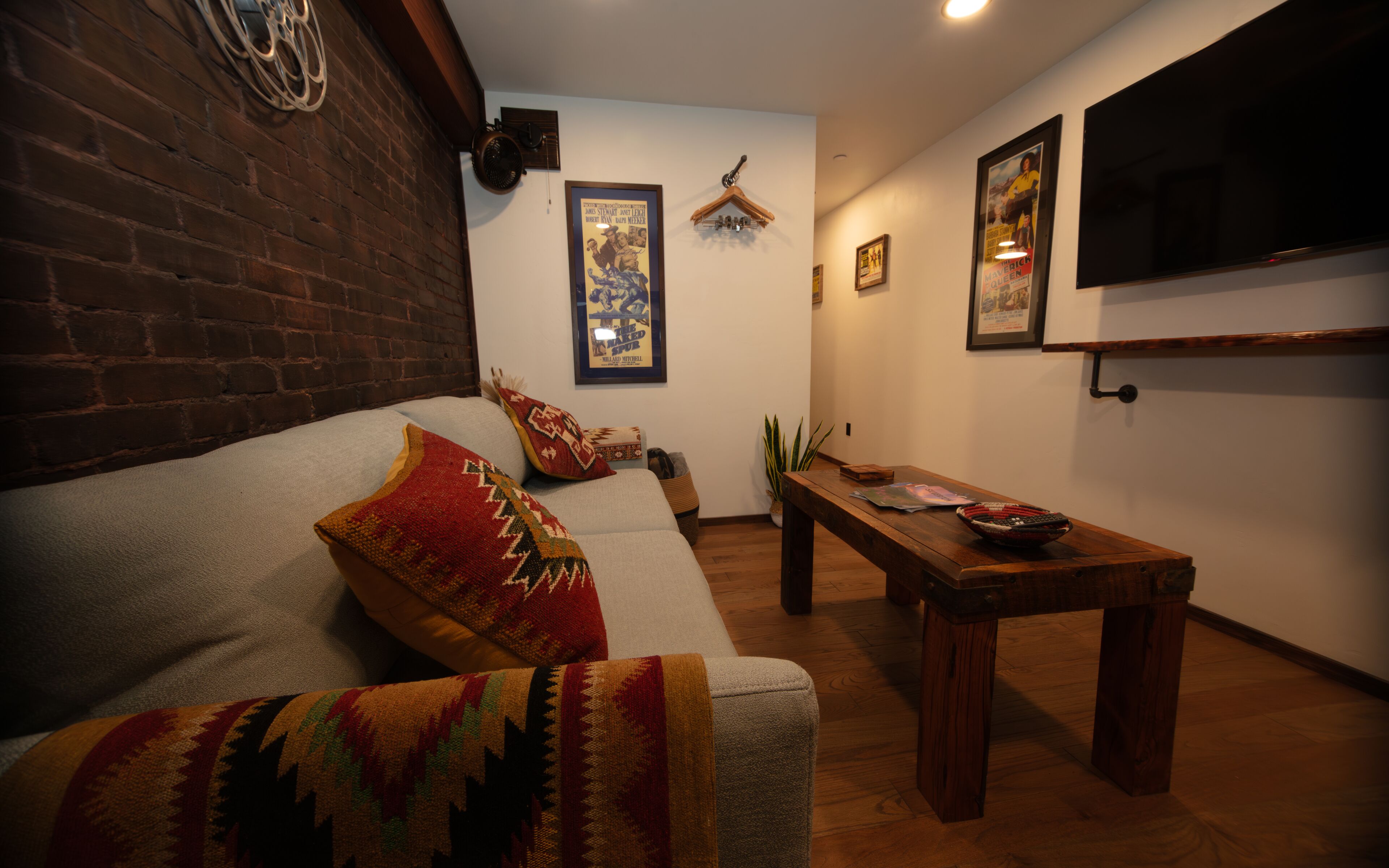 Una sala de estar acogedora con pared de ladrillo, un sofá con cojines estampados, una mesa de centro de madera y un televisor montado en la pared.