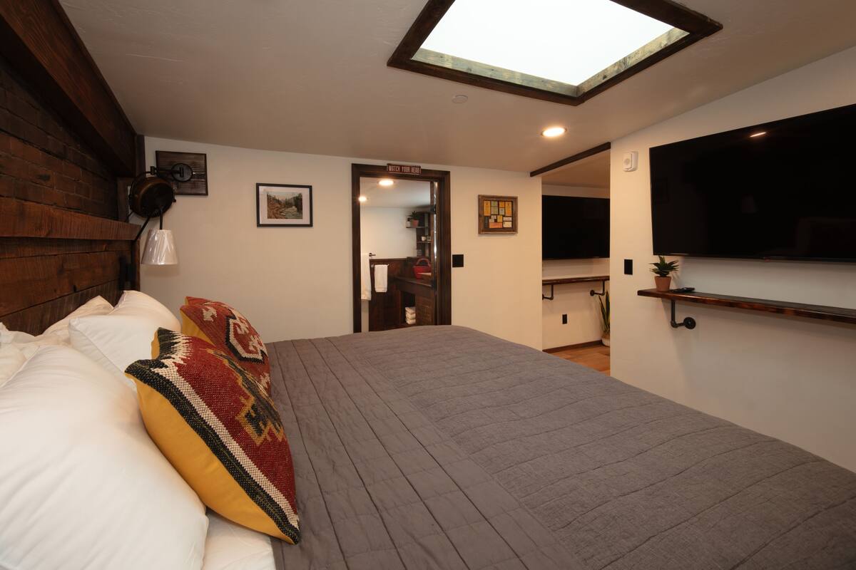 A bedroom with a skylight, a bed with pillows, a bedside table, a mirror, and a TV mounted on the wall.