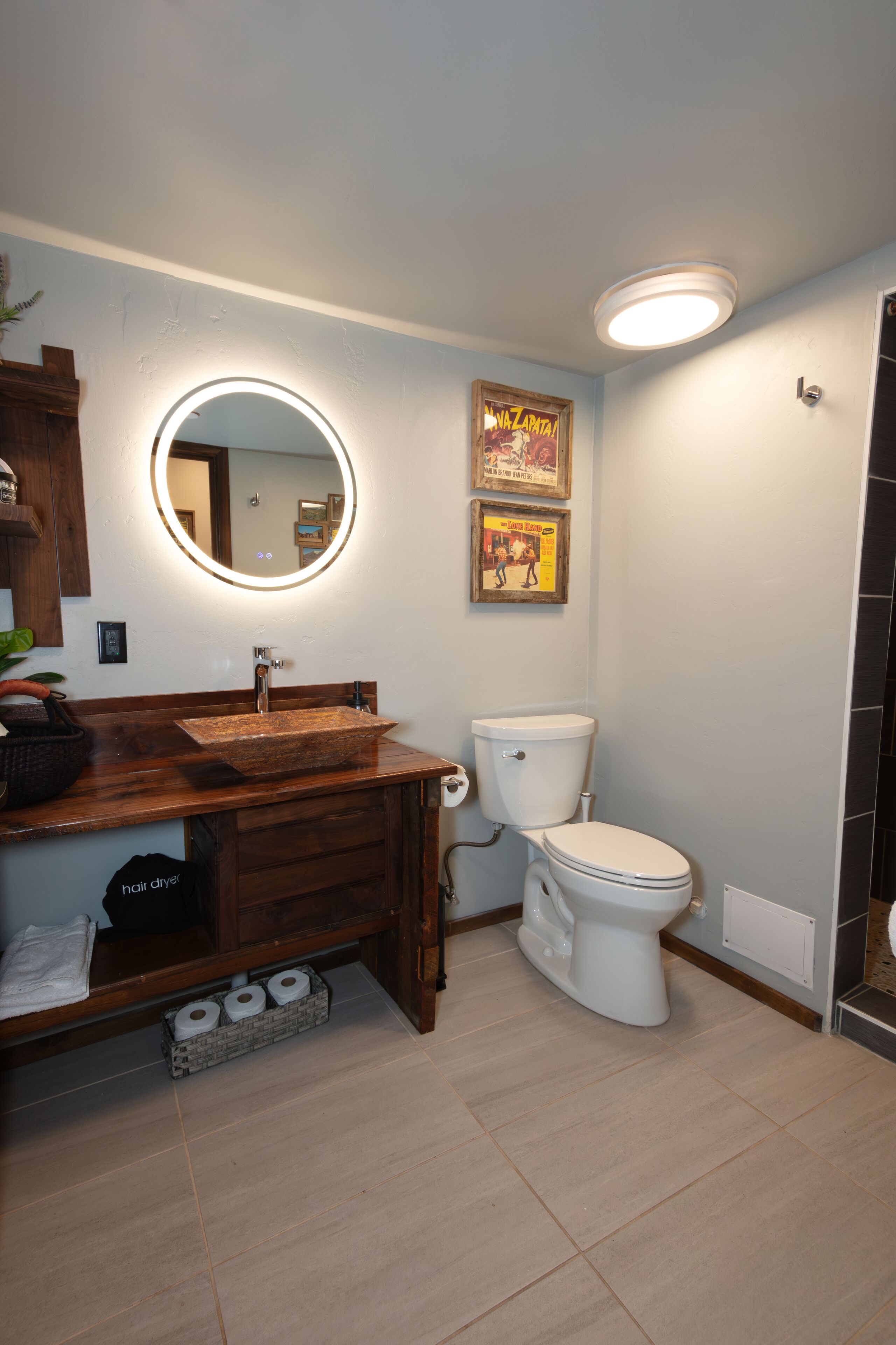 A bathroom with a wooden vanity, a round mirror with lighting, a toilet, and two framed pictures on the wall.