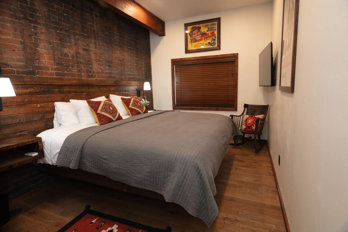 A bedroom with a brick wall, a wooden headboard, a large bed with a gray quilt, a wooden nightstand with a lamp, and a framed picture on the wall.