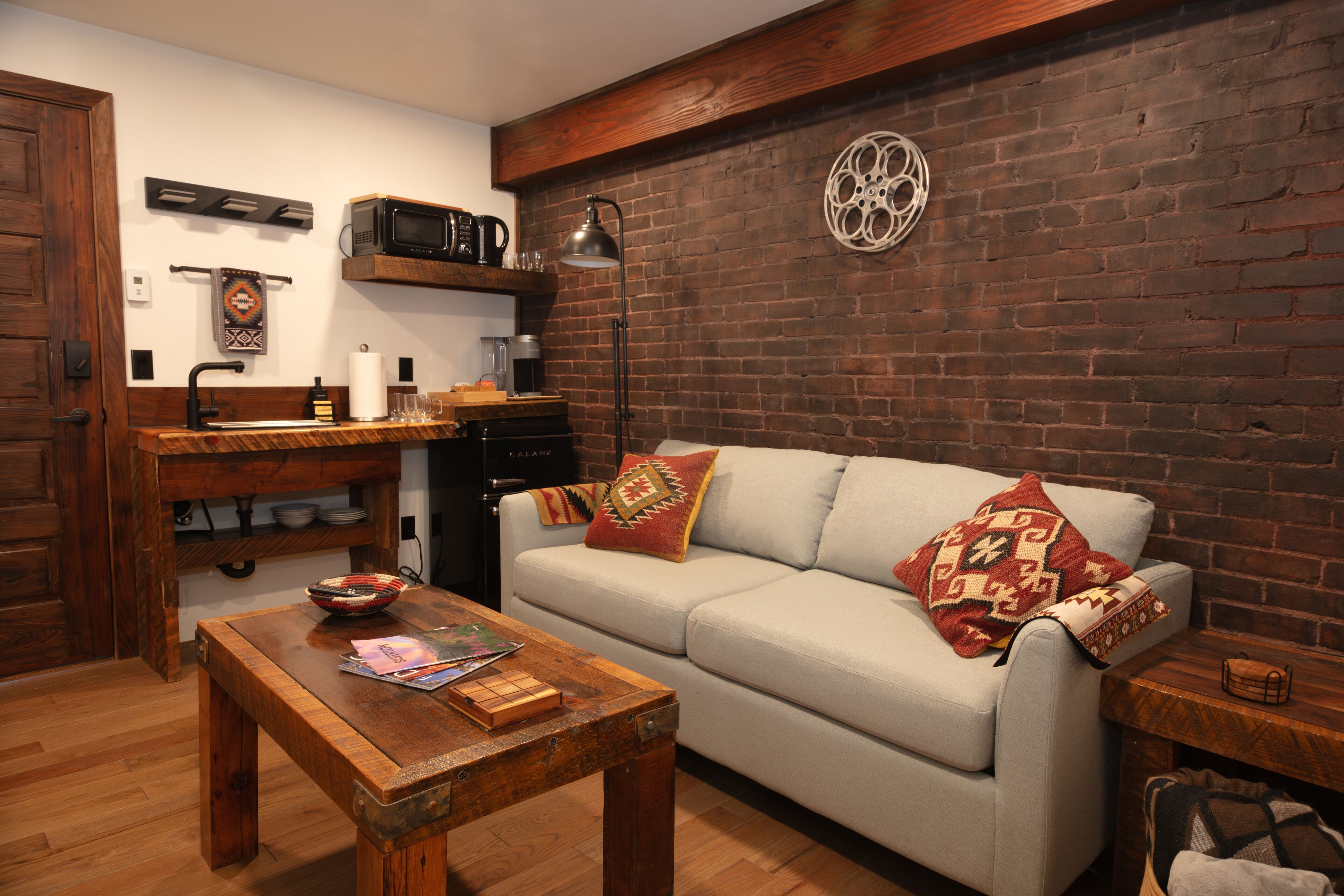 A cozy living room with a wooden coffee table, a sofa with patterned cushions, a brick wall, and a small side table.