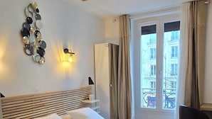 Et moderne soveværelse med en stor seng, trægulv, en vægmonteret lampe og udsigt til bygninger gennem et vindue med gardiner.