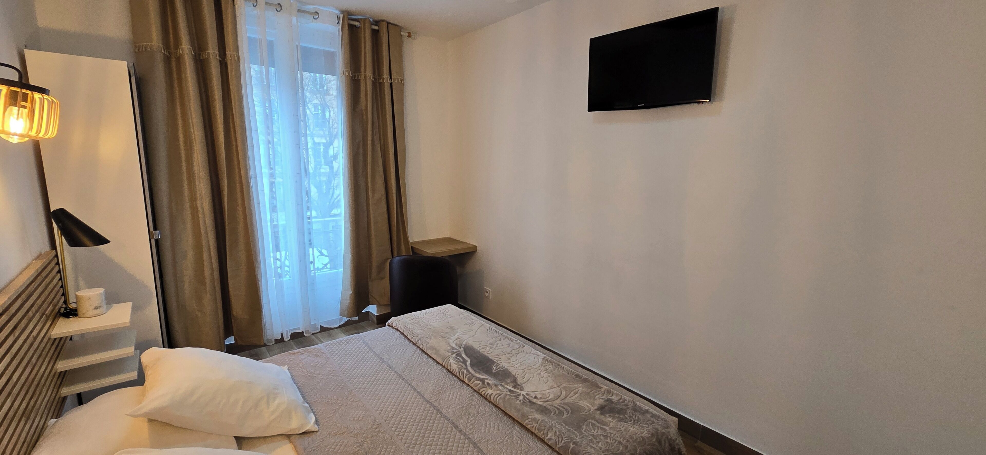 A hotel room with a bed, a bedside table with a lamp, a flat-screen TV mounted on the wall, and a window with curtains.