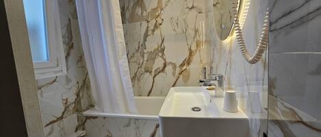 A bathroom with a marble wall, a white sink, a glass shower enclosure, and a mirror with lights.