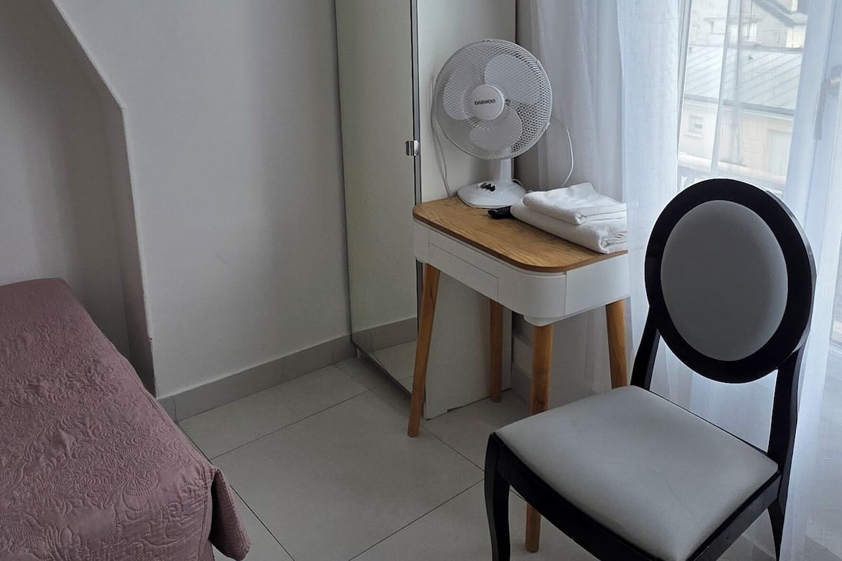 Une petite chambre moderne, équipée d’un lit, d’une chaise, d’un bureau et d’un ventilateur sur pied.