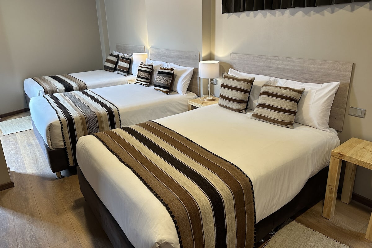 Habitación de hotel con dos camas, cada una con ropa de cama a rayas y varias almohadas. Hay una mesita de noche con una lámpara y un pequeño mueble de madera.