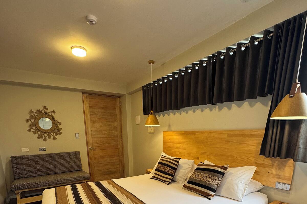 Un dormitorio con una cama grande, cabecera de madera, una banca, una mesita de noche y una lámpara de pared.
