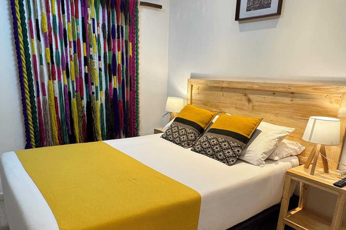 Una cama bien tendida con una manta amarilla, un cabecero de madera y almohadas estampadas. Se observa una mesita de noche con una lámpara y un cuadro en la pared.