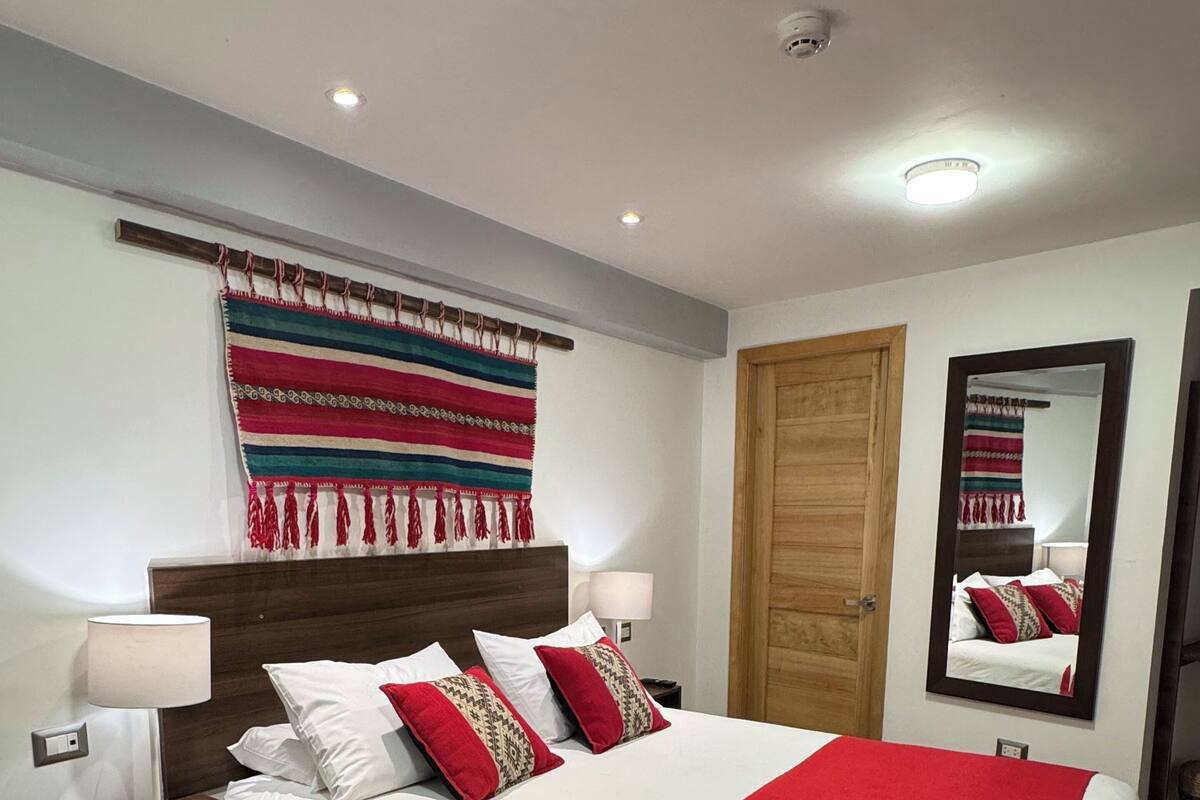 Un dormitorio ordenado con una cama grande, cabecera de madera, mesita de noche con lámpara, espejo en la pared y un tapiz colorido.