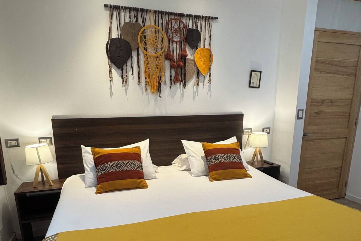 Una cama bien hecha con un edredón amarillo, dos lámparas de noche y una decoración en la pared sobre la cama.