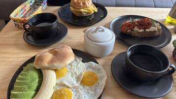 Uma mesa de madeira com um prato de ovos, abacate, banana, uma xícara de café, um bule e um sanduíche.