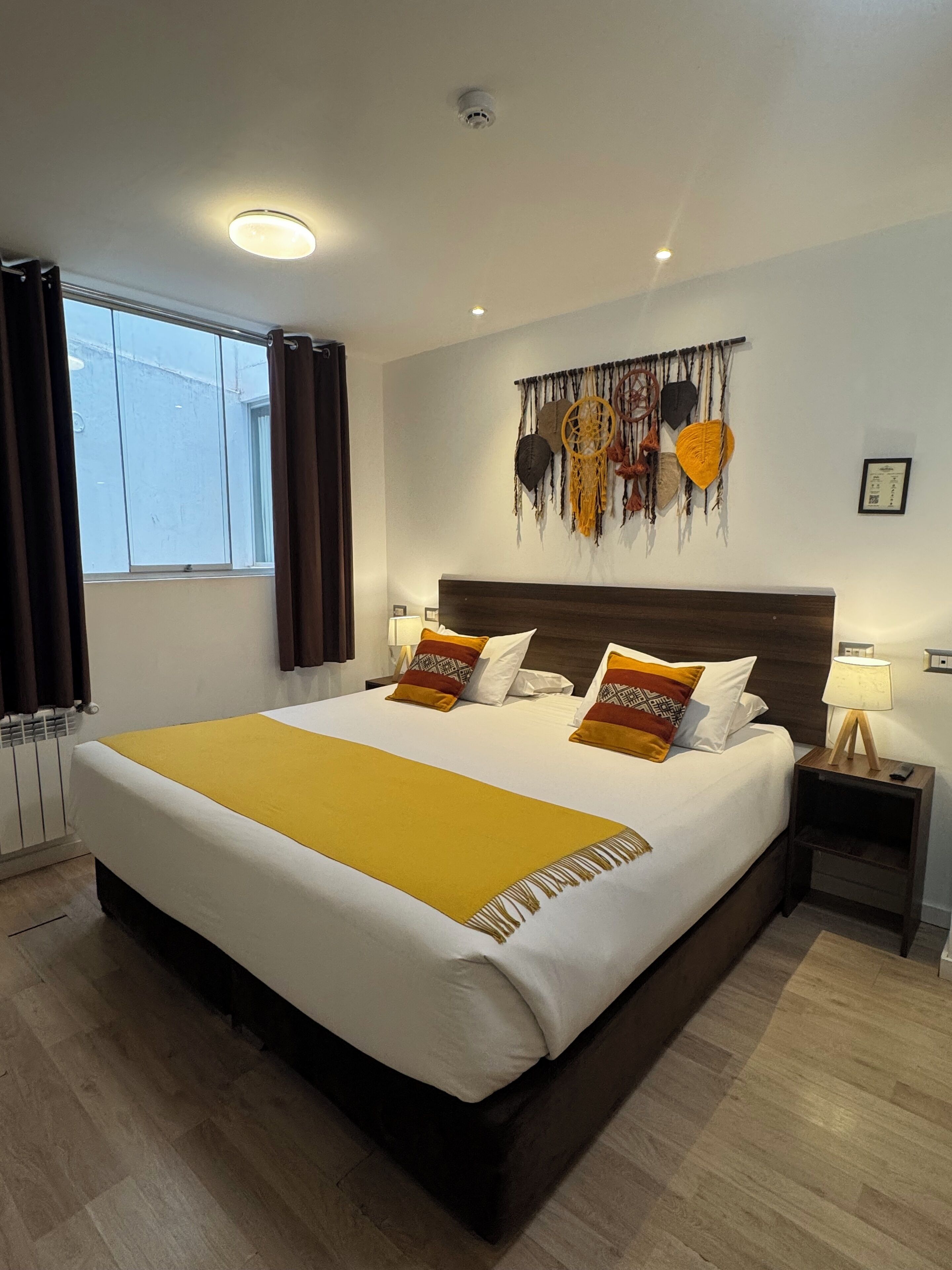 A large bed with a yellow throw blanket, a dark headboard, and patterned pillows. There is a wall-mounted dreamcatcher decoration and a small bedside table with a lamp.