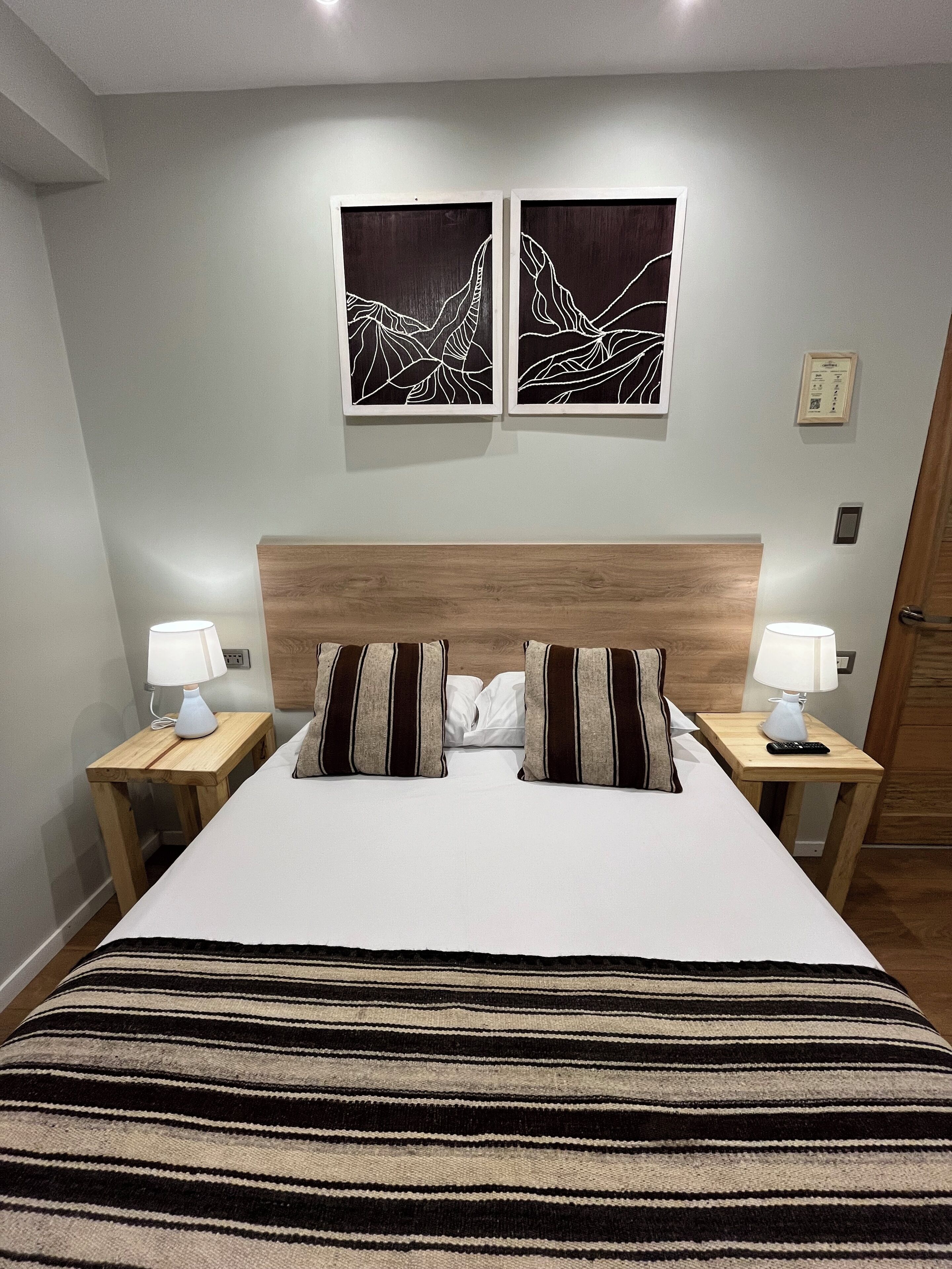 A neatly made bed with striped bedding, two bedside tables with lamps, and two wall-mounted artworks depicting abstract mountain designs.