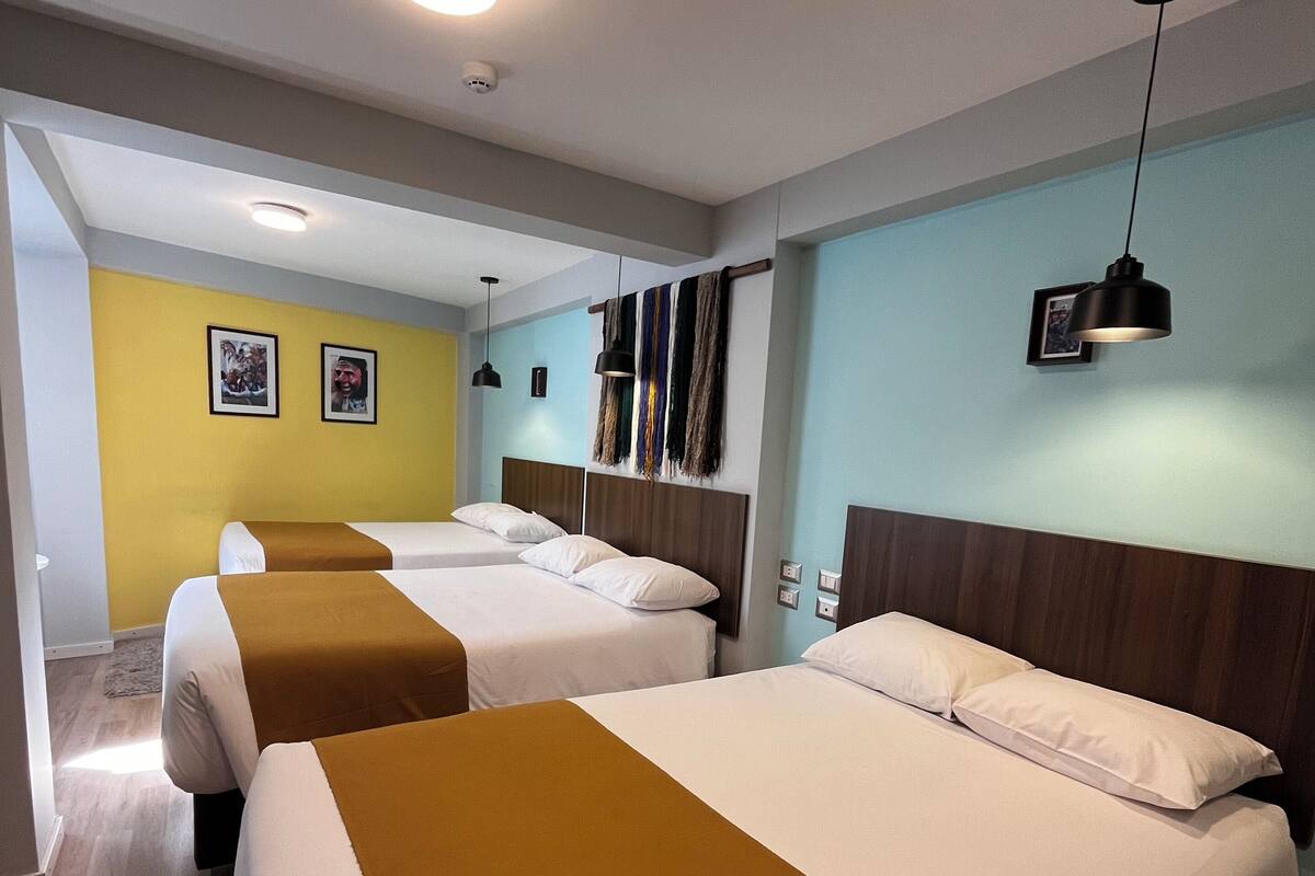 Habitación de hotel con tres camas, cabecera de madera, pared de acento amarilla y un tapiz colgante.