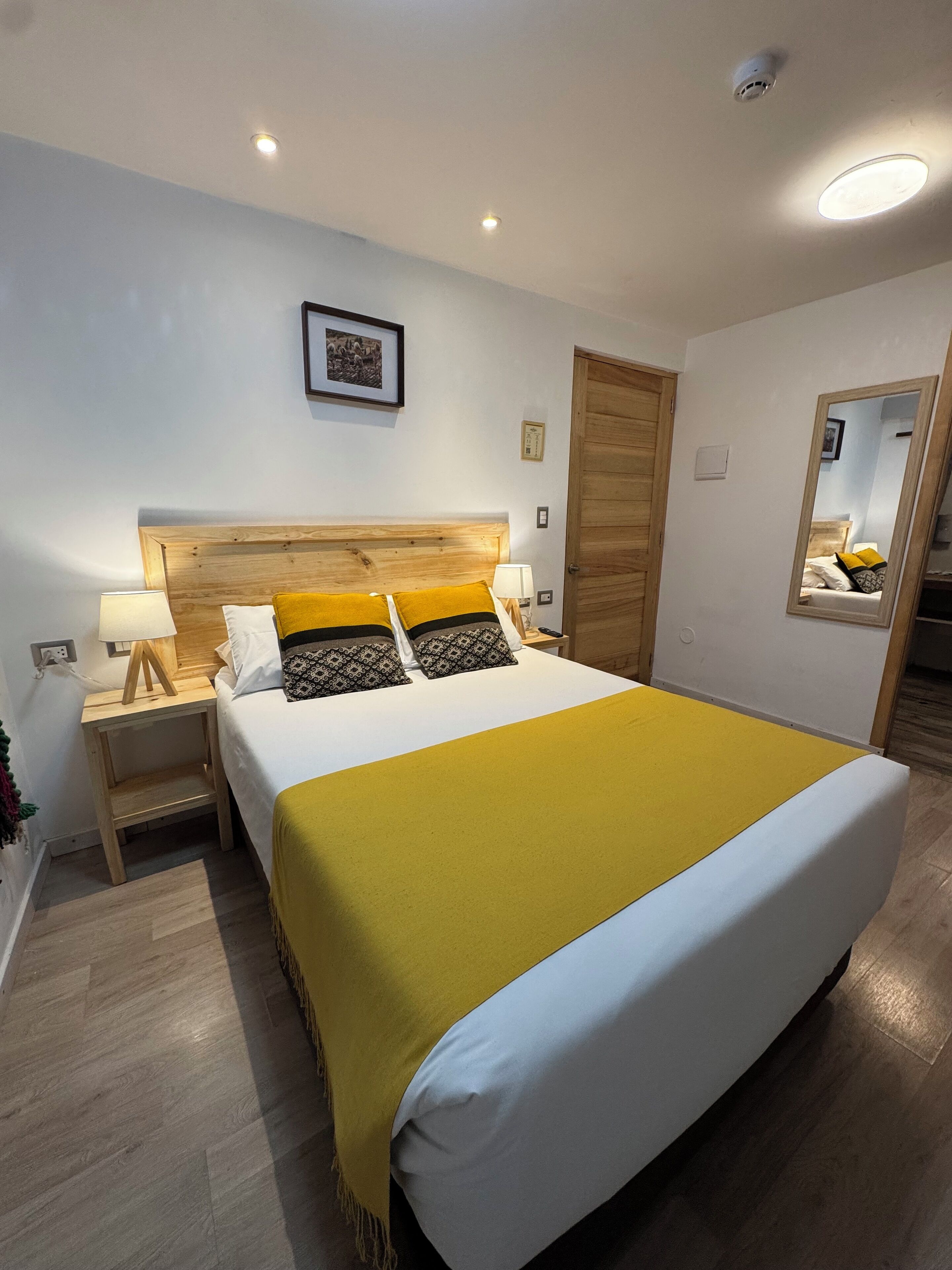 A neatly arranged bedroom with a wooden headboard, a bed with a yellow blanket, a bedside table with a lamp, and a mirror on the wall.