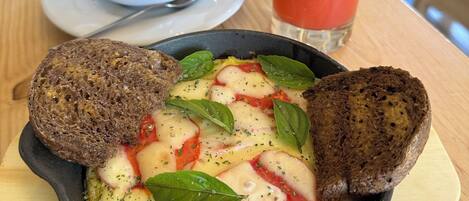 A breakfast setting with a bowl of eggs, tomatoes, and basil, accompanied by slices of bread and a glass of red juice.