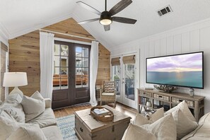 A cozy living room with a large TV, wooden walls, a ceiling fan, and a sliding glass door leading to a balcony. - Little Blue Heaven 30A Beachside Cottage w/ Pool (Santa Rosa Beach)