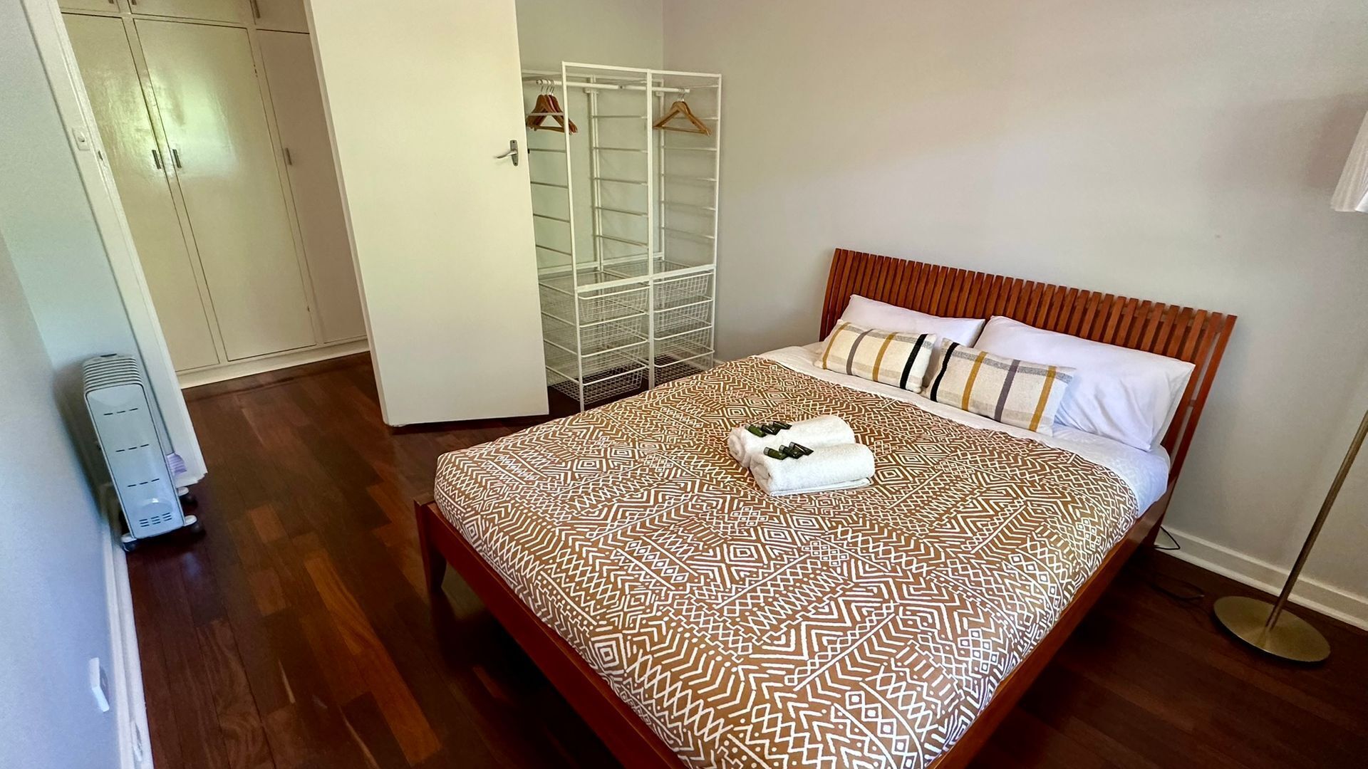 A neatly arranged bedroom with a patterned bedspread, a wooden headboard, and a bedside table with a towel and bottles.
