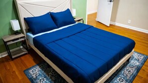 A bed with a white headboard and blue bedding, a nightstand with a lamp, and a patterned rug on a wooden floor. - Warm spacious midsize home w/ comfort and fun (Glendale)