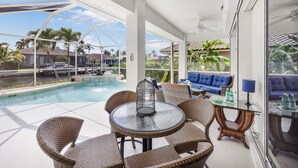 A covered outdoor area with a pool, wicker furniture, and a glass table.