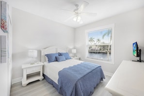 Une chambre à coucher comprenant un lit, des tables de chevet, un bureau, un ventilateur de plafond et une fenêtre donnant sur des palmiers et un quai.