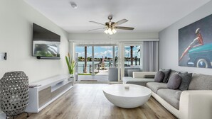 A modern living room with a large flat-screen TV, a ceiling fan, a white coffee table, and a sofa with grey cushions.