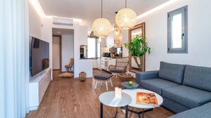 Una sala de estar moderna con un sofá gris, una mesa de centro y sillas de mimbre. Hay un televisor de pantalla plana montado en la pared, una planta en maceta y una ventana con vista a la cocina.