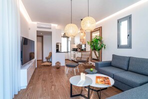Un salon moderne avec un canapé gris, une table basse et des fauteuils en osier. Un téléviseur à écran plat est fixé au mur, ainsi qu'une plante en pot et une fenêtre donnant sur la cuisine.