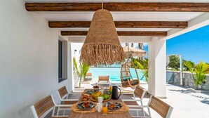 Un espace de restauration en plein air avec une table en bois, des chaises blanches et un luminaire suspendu en fibres tressées.