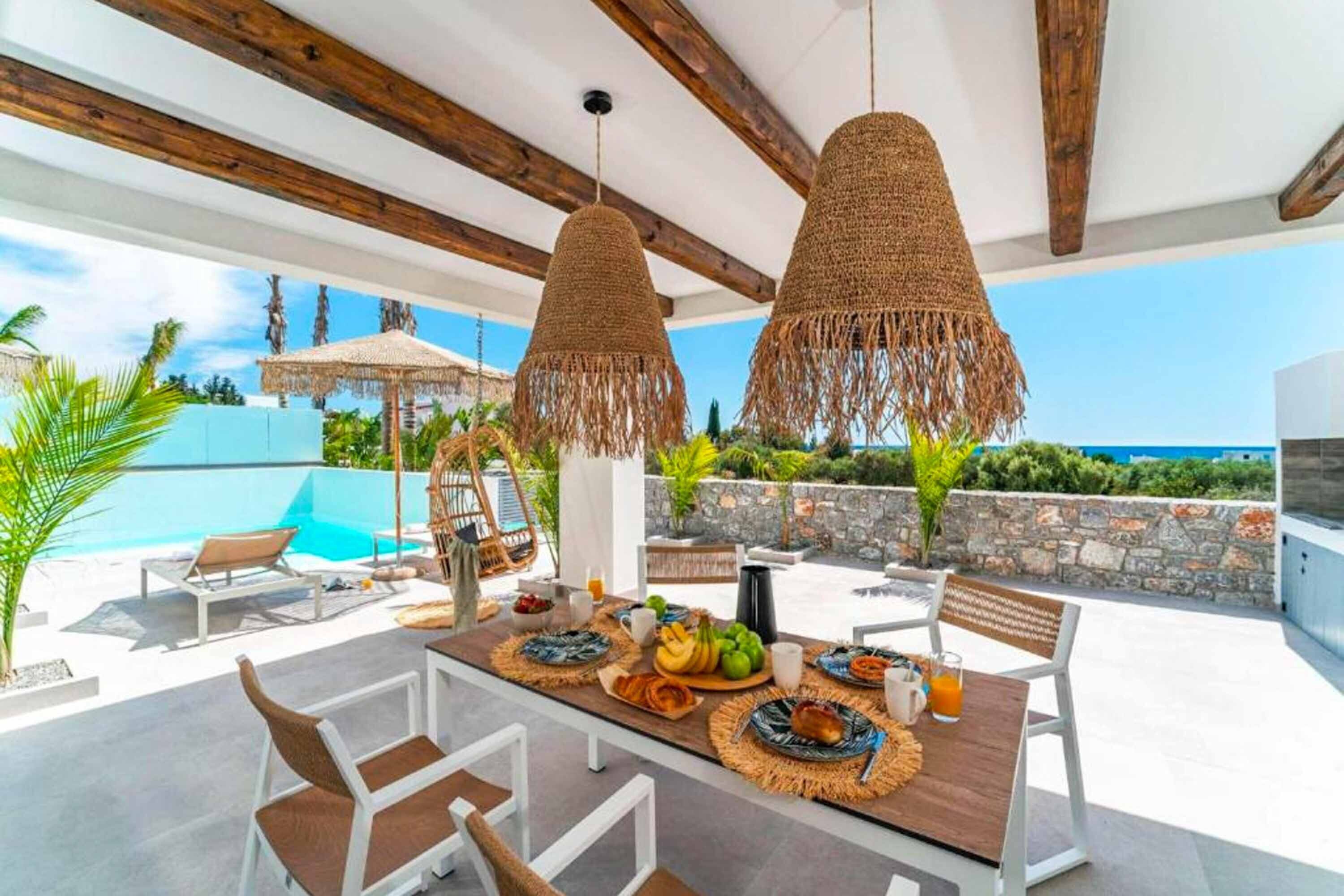 Un área de comedor al aire libre con una mesa lista para el desayuno, rodeada de sillas y con vistas a una piscina y follaje.