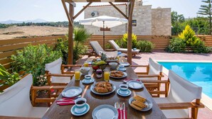 Uma área de refeições ao ar livre com vista para a piscina, móveis de madeira e uma mesa posta para o café da manhã.