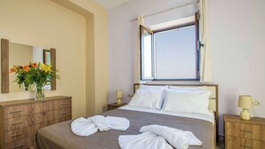 A hotel room with a bed, a wooden dresser, a nightstand, a window with curtains, and a vase with flowers. - Villa Ourania Klio - Two Bedroom Villa, Sleeps 4 (Crete)