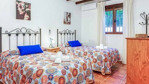 A bedroom with two beds, each with a patterned bedspread and blue pillows. There is a wooden nightstand with a lamp and a decorative item. A window with curtains is visible, and a wooden cabinet is in the corner.